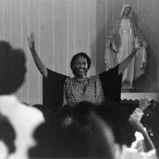 Sister Thea Bowman with arms upraised and in African attire before a congregation